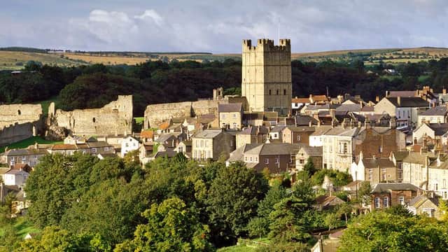 Richmond Castle rises above a river, framed by lush greenery and a stone bridge, under a blue sky with scattered clouds.