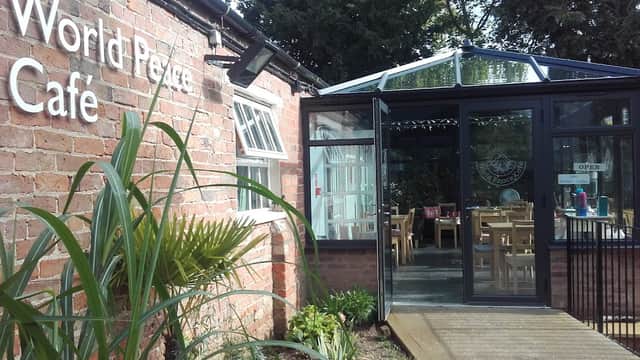 Entrance to World Peace Cafe with glass conservatory, surrounded by greenery and rustic brick walls.