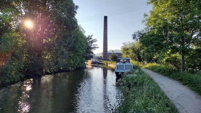 A serene canal scene in Skipton, Yorkshire, with a boat, lush greenery, and a tall brick chimney in the background.