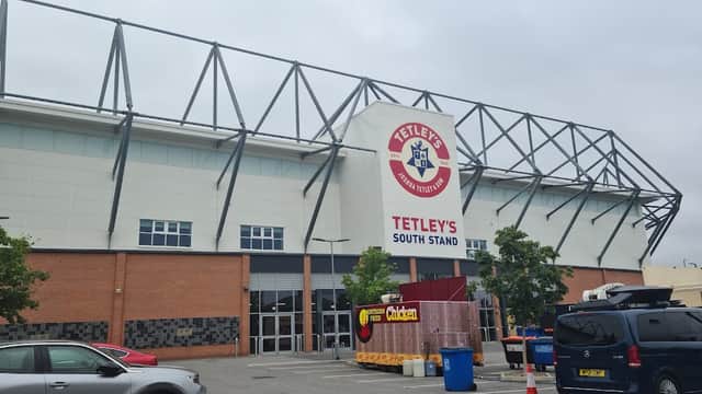 Tetley's South Stand building with a car park in front, featuring parked cars and nearby trees under a cloudy sky.