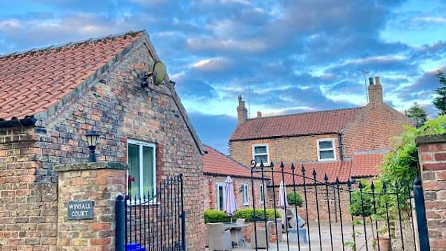 Rustic brick buildings with a gated entrance, under a blue sky with clouds, in a tranquil countryside setting.