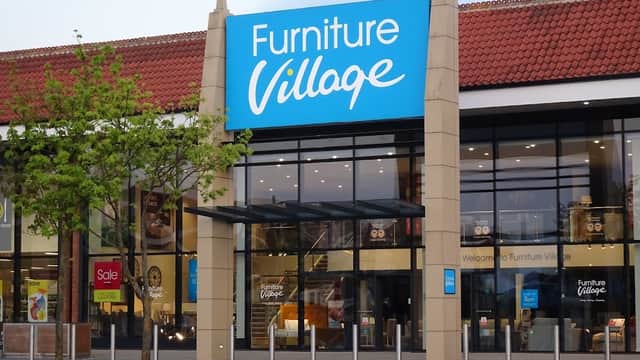 Bright blue sign reading "Furniture Village" on a modern storefront with large glass windows and a tree in front.
