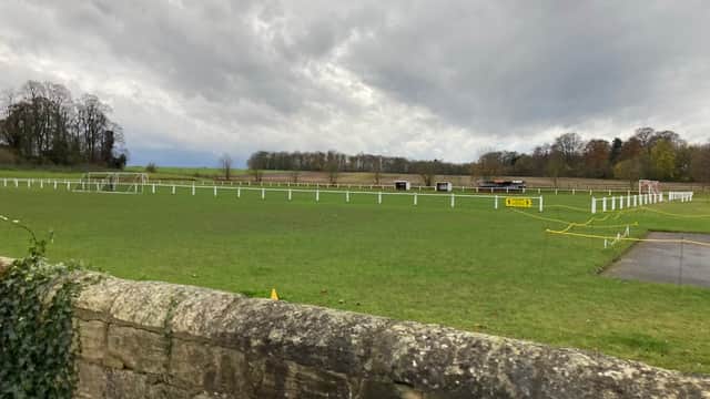 Green football pitch with white goalposts, surrounded by a stone wall and trees under a cloudy sky.
