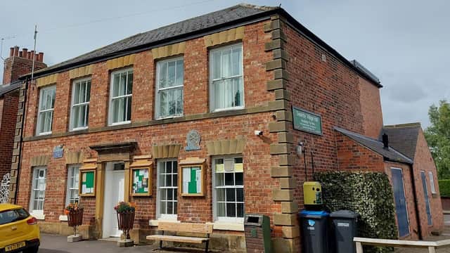 Red brick building with large windows, a bench outside, and signs for Swainby Village Hall. Green bins nearby.