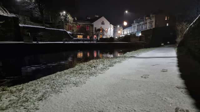 Snow-covered path beside a canal at night, with illuminated buildings and trees in the background.