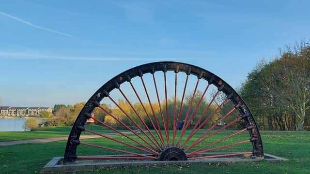 Wath Main Pit Head Winding Wheel stands prominently against a clear blue sky, surrounded by greenery and a lake.