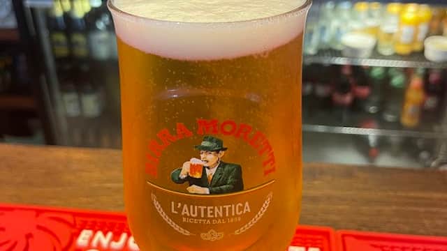 A pint of beer in a branded glass sits on a bar mat, with a blurred background of bottles and bar equipment.