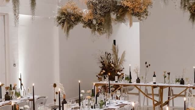 Elegant floral arrangements hang overhead in a bright venue, with long tables set for a celebration, featuring candles and wine.