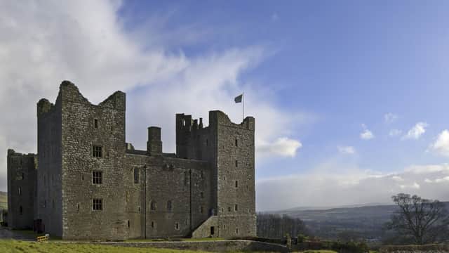 Historic Bolton Castle features stone walls, towers, a flag, and lush green surroundings under a bright blue sky.