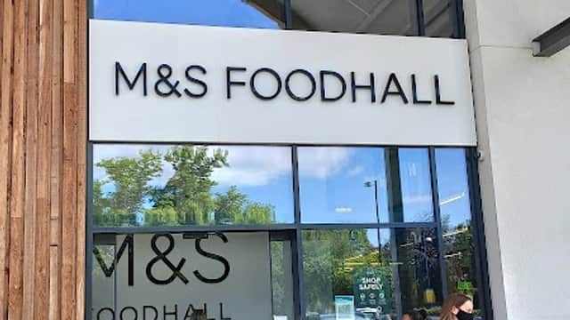 Modern storefront of M&S Foodhall with large glass entrance, wooden accents, and shoppers outside.