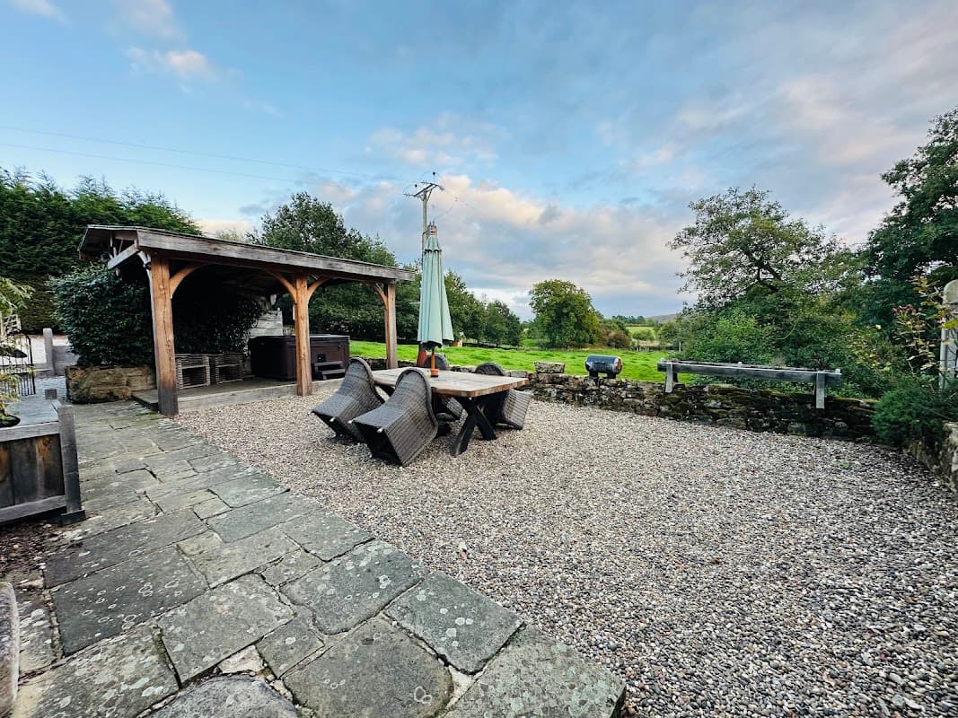 Outdoor seating area with a wooden pergola, gravel surface, and lush greenery in the background.