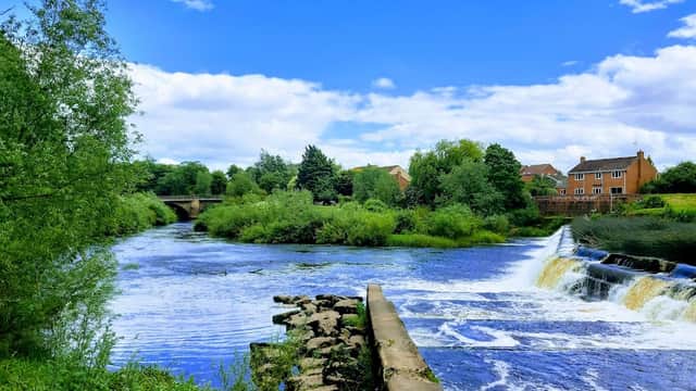 Scenic river view with lush greenery, a waterfall, and houses under a blue sky with fluffy clouds in Boroughbridge.