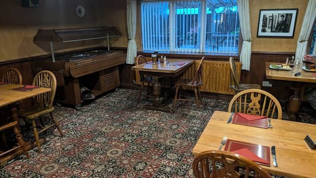 Cozy dining area with wooden tables, chairs, a patterned carpet, and a large window with sheer curtains.