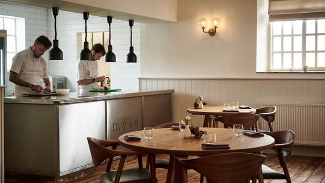 Modern restaurant interior with wooden tables, stylish lighting, and chefs preparing food in an open kitchen.