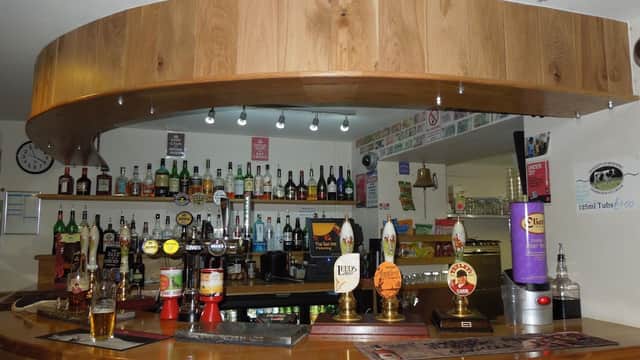Wooden bar with various bottles and taps, glasses, and a clock on the wall in a cozy pub setting.