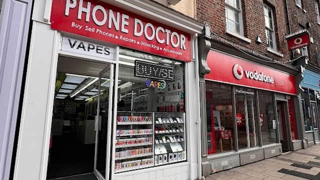 "Storefront of Phone Doctor with signage, displaying phones and vapes, next to a Vodafone store in York."