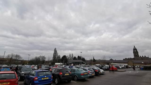 Busy car park filled with various cars under a cloudy sky, with a clock tower visible in the background.