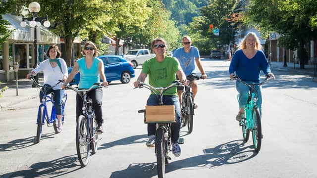 A group of five people riding bicycles on a sunny street, surrounded by trees and shops.