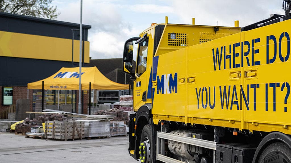 Yellow truck with "WHERE DO YOU WANT IT?" parked outside MKM Building Supplies, featuring a tent and building materials.