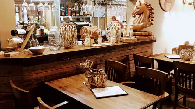 Rustic restaurant interior featuring wooden tables, a bar with decorative jars, and a wooden horse sculpture.