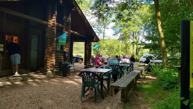 Log cabin cafe surrounded by trees, with patrons seated at outdoor tables and a gravel path leading to the entrance.