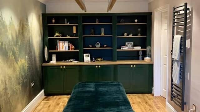 A serene therapy room featuring a massage table, wooden shelving, and calming forest mural on the wall.