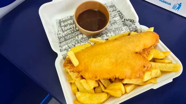 Crispy battered fish on a bed of golden chips, served with a small cup of brown sauce in a takeout container.