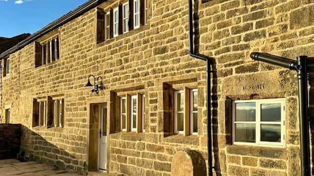 Stone cottage with multiple windows, set against a blue sky, featuring a paved area and stone wall in East Morton, Yorkshire.