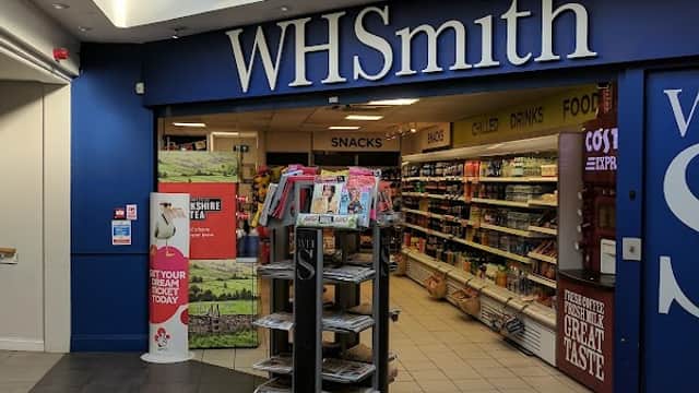 WHSmith storefront with blue signage, shelves stocked with snacks and drinks, and a display stand for magazines.