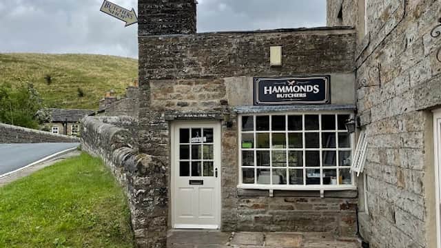 Hammonds Butchers shop with stone facade, large window, and sign, set against a green hillside in Bainbridge, Yorkshire.