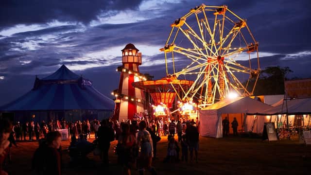 Colorful festival scene at dusk with a ferris wheel, tents, and a lighthouse structure, filled with people enjoying the event.