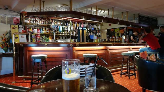 Wooden bar with various drinks, bar stools, and tables; patrons chatting in a cozy, warmly lit atmosphere.