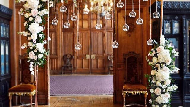 Floral arch adorned with white roses and hanging glass orbs, set in an elegant interior with wooden paneling.