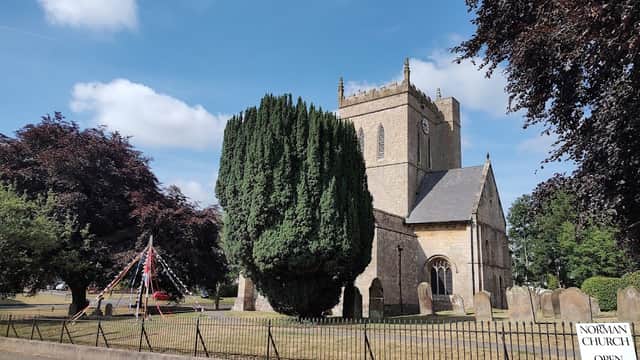 Norman church with tall trees, gravestones, and a sign reading "Open for Coffee" against a blue sky.