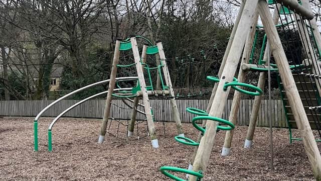 Playground equipment with climbing structures and slides, surrounded by wood chips and bare trees in Holme Valley.