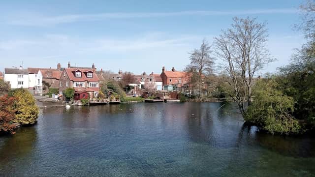 Scenic view of Nafferton Mere with clear water, surrounded by quaint houses and trees under a blue sky.