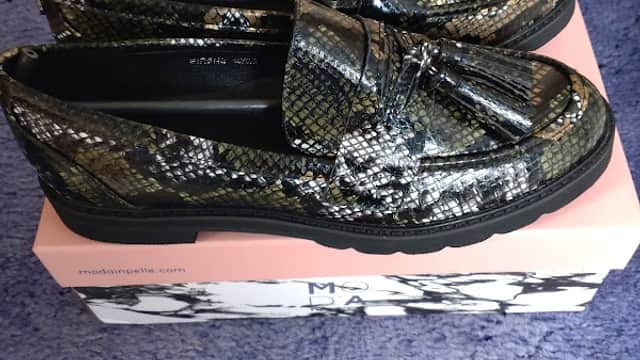 Stylish black and silver snake-print loafers on a pink shoe box, set against a plush purple background.