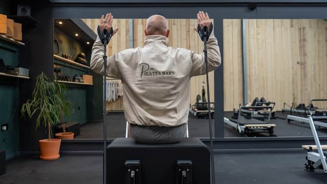 A person in a Pilates jacket sits on a reformer, facing a mirror in a well-lit studio with exercise equipment.