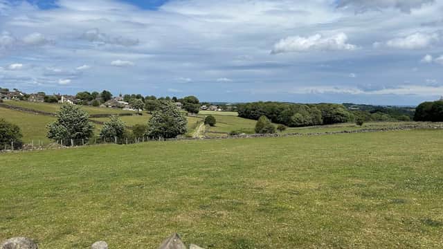 Lush green fields and stone walls under a blue sky, with distant village houses and trees in Upper Cumberworth, Yorkshire.