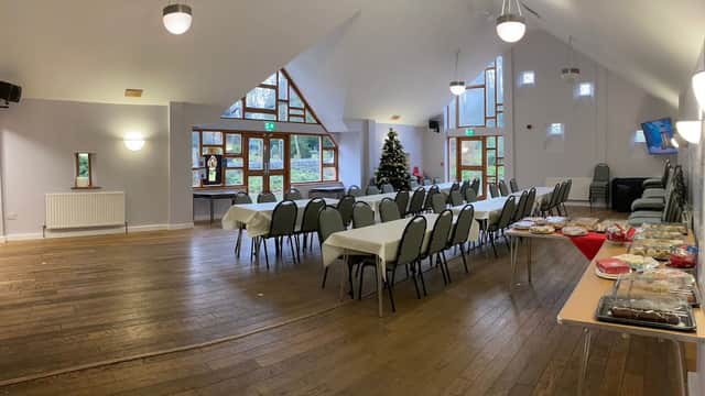 Bright hall with wooden floors, tables set for an event, a Christmas tree, and large windows letting in natural light.
