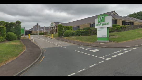 Entrance to Holiday Inn with green signage, surrounded by neatly trimmed hedges and a cloudy sky in Brighouse, Yorkshire.