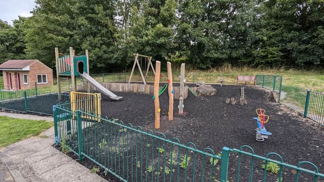 Playground with a slide, climbing structure, swings, and benches surrounded by a green fence and trees in the background.