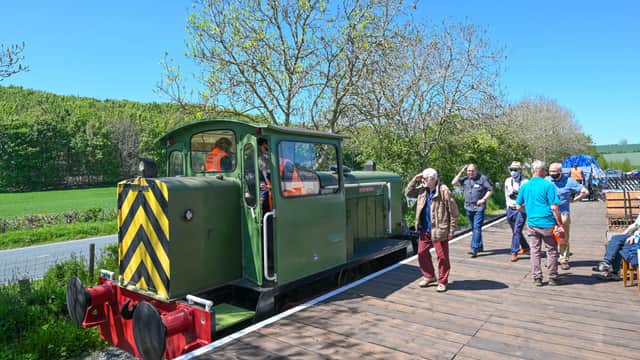 A green locomotive at Yorkshire Wolds Railway with visitors enjoying the sunny day and lush green fields in the background.