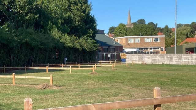 Green field with wooden fences, buildings in the background, and a church spire visible against a clear blue sky.