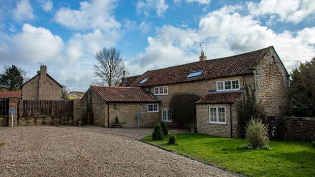 Stone cottages with a red-tiled roof, surrounded by green grass and gravel, under a partly cloudy sky.