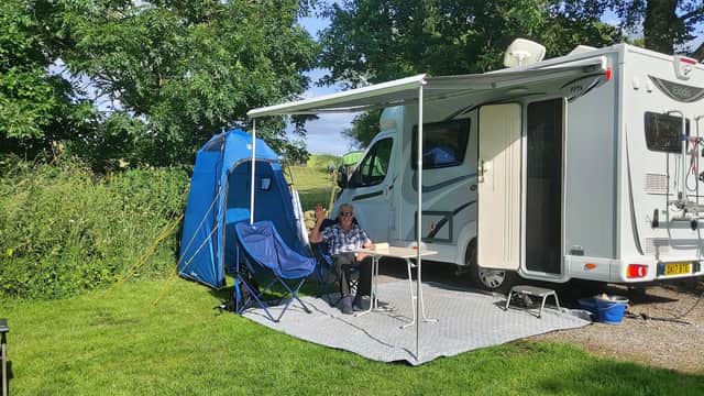 A camper van with a blue tent nearby, set on green grass, with a person sitting at a table under a canopy.