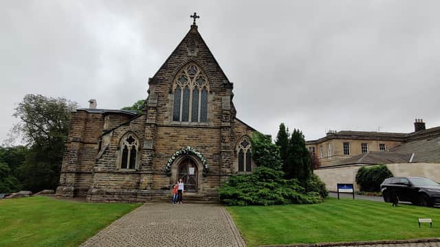 Victorian-style church with stone faΓ§ade, arched windows, and a cross atop, surrounded by greenery and a gravel path.