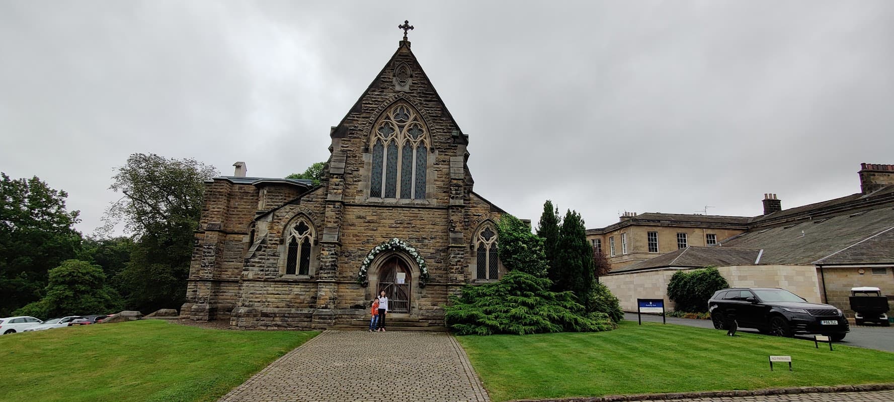 Victorian-style church with stone façade, arched windows, and a cross atop, surrounded by greenery and a gravel path.