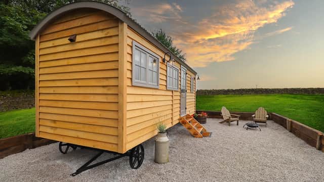 Yellow wooden hut on a gravel campsite, with a fire pit and Adirondack chairs, set against a sunset sky.