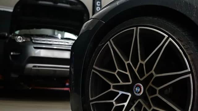 Close-up of a stylish alloy wheel with a blurred car in the background and another vehicle with an open hood.